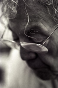 This black & white closeup portrait photograph is an artistic shallow-focus impression of Hans Erni at work in his studio. The view is on his left temple from an approximately 45 degree angle between front and side. His head is tilted forward and he is looking down. In focus are primarily a big curl hanging over his forehead, a narrow long strand of gradually curling hair extending even beyond his left eyebrow, and the top left corner of his glasses.