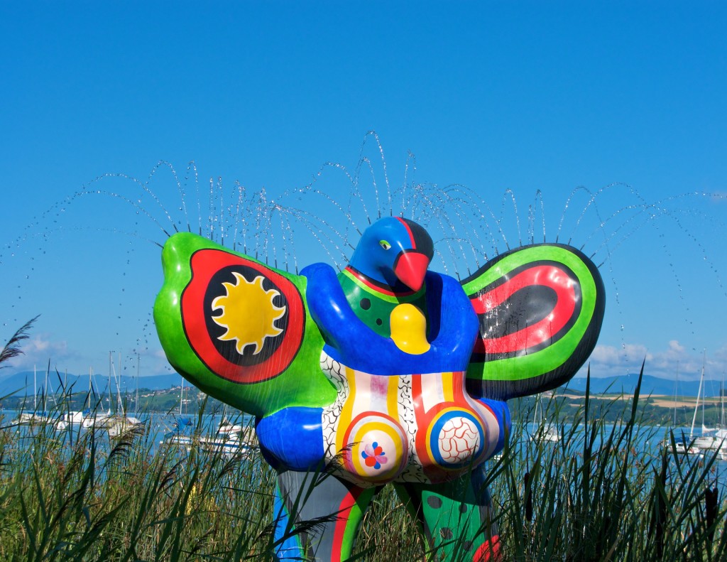 Color photograph of the Nana fountain/sculpture, which stands in an expanse of reeds with anchored sailboats in the background. The huge, colorful sculpture of a voluptuous woman/angel/bird has water jets along the upper edge of its wings and on top of its bird head. Lime green, royal blue, and fire-engine red colors dominate on the glossy surface of the huge polyester sculpture.