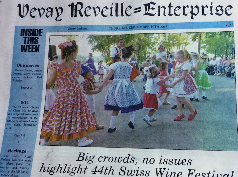 Detail from a Vevay Reveille-Enterprise cover, showing the masthead and a color photograph of costumed adults and children in the city park with the Ohio River as a backdrop. The children are dancing in pairs holding hands while onlookers are seated on the fringe. The headline below reads: Big crowds, no issues highlight the 44th Swiss Wine Festival.