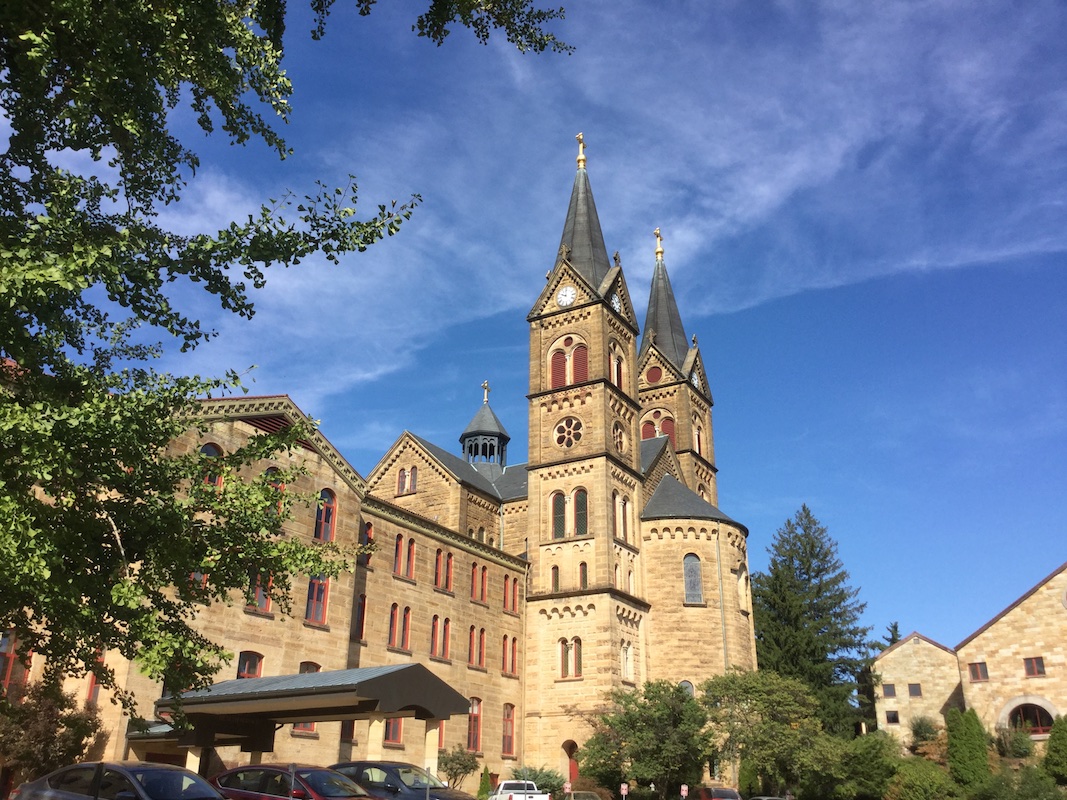 Color photograph of the Saint Meinrad Archabbey church at the center with the two main towers, big apse between them at the east end, and the small crossing tower visible in the back. To the left is St. Anselm Hall of the Seminary & School of Theology with the long canopy of the Health Services entrance and some cars in the foreground. At right, detached from the church, is the south-west corner of the Monastery.