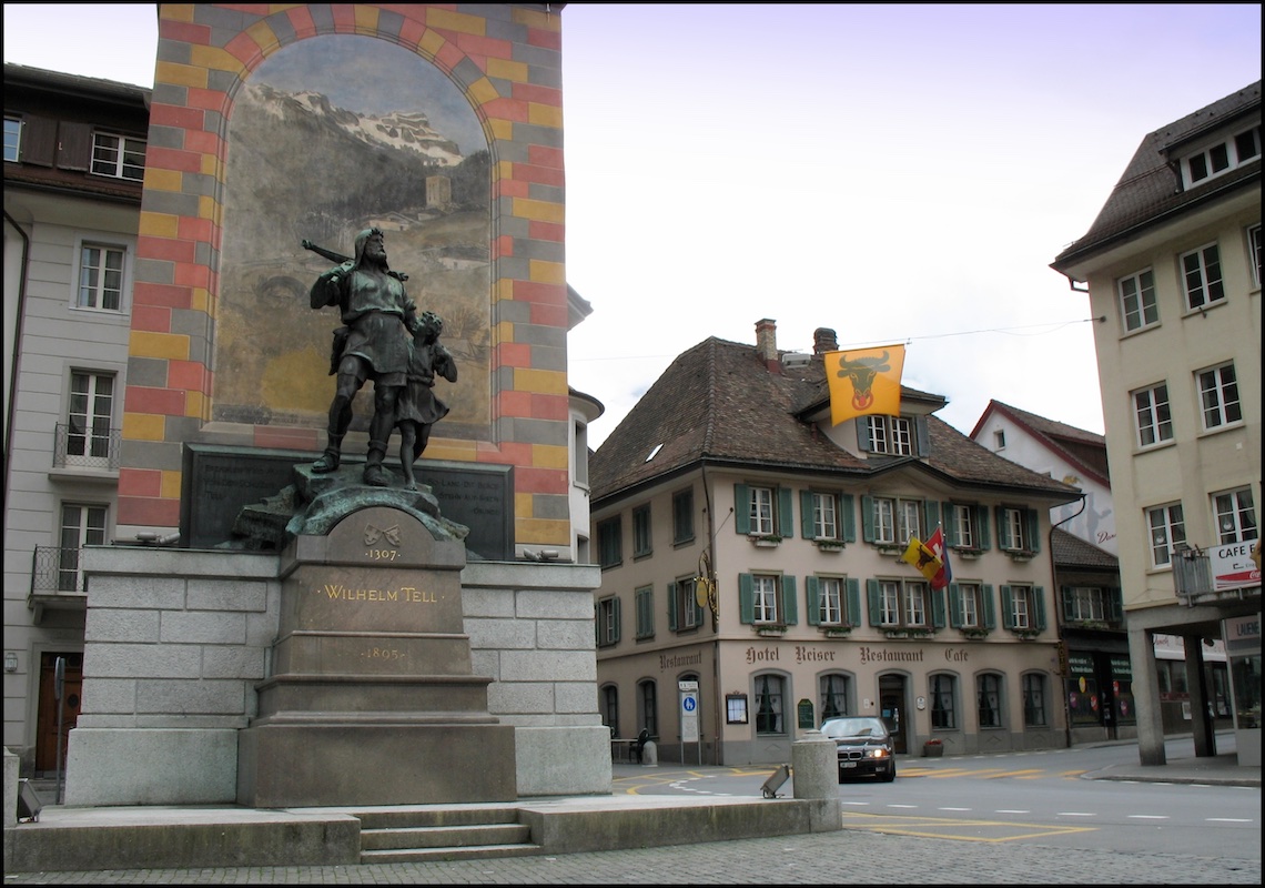 Color photograph of the Wilhelm Tell monument in Altdorf, Switzerland. The bronze statue is displayed in front of a painting of a