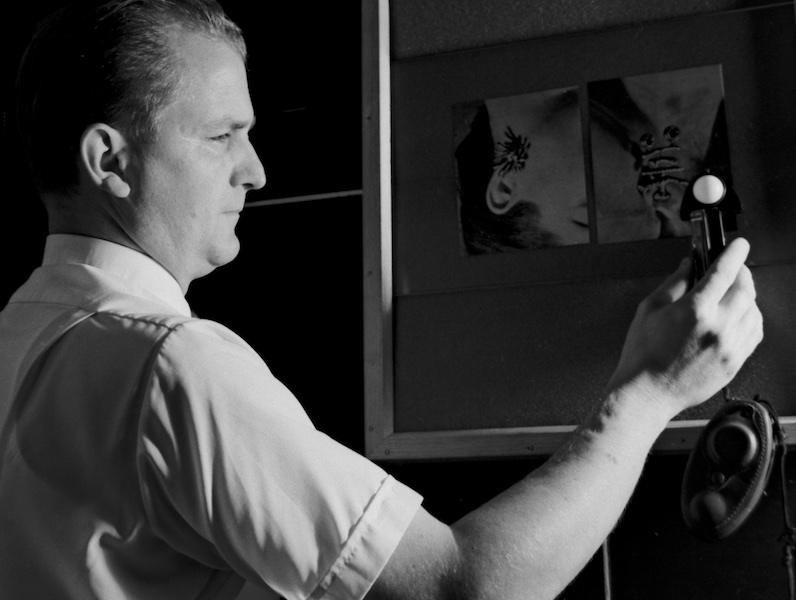 Black and white photograph showing a right head and chest profile view of James Perret wearing a short-sleeved white shirt in a dark room. He is watching an incident-light meter that he hold in front of a framed pasteboard where one can barely make out a couple of attached photographs. Directional lighting from the right dramatically emphasizes his silhouette, right side of face and right ear as well as his angled right arm and the white semi-spherical sensor of the meter he is holding up.