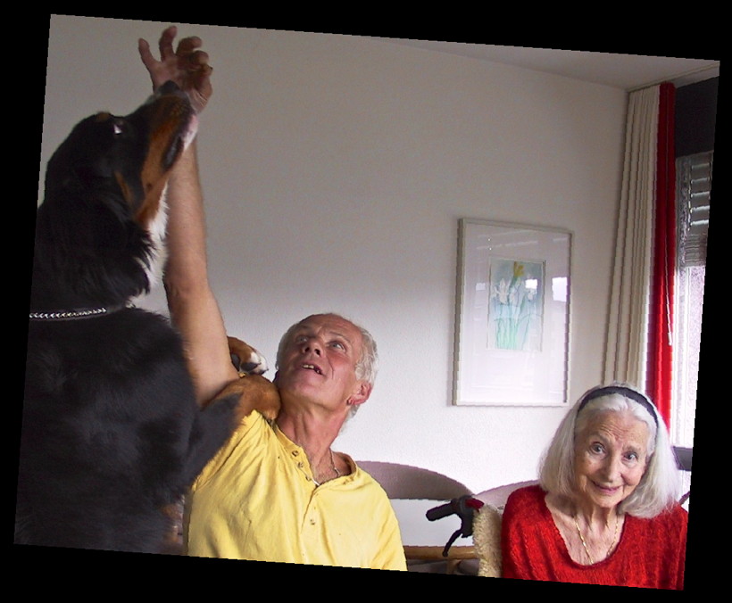 Color photograph of a Bernese mountain dog, a sitting middle-aged man in a yellow short-sleeve shirt, and an elderly woman with white hair and a red sweater, in a wheelchair. The man has his right arm stretched up as high as possible, holding a treat between his fingers. The dog is standing up, with her front legs wrapped around both sides of the man's arm and resting her paws on his shoulder, stretching her head toward the treat. The dog's and the man's eyes are focused on the treat; the woman is looking straight into the camera.