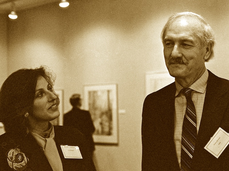 Sepia-toned black and white photograph of a dark-haired woman looking up to a tall, white-haired man who could probably rest his chin on the top of her head if they were standing much closer together. Visible in the soft-focused background between them is an art gallery wall with a person looking at one of the pictures.