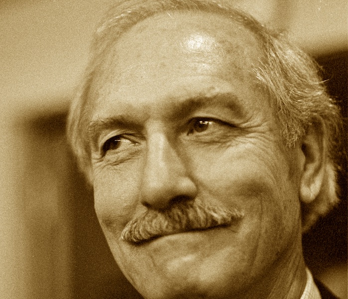 Sepia-toned black and white portrait of a man with a mustache, lively eyes, and a broad smile.