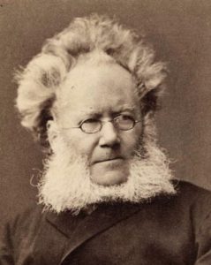 Sepia-toned black and white portrait photograph of a man with unruly hair (even more reminiscent of Albert Einstein's) and a square white beard spreading out from the cheeks and below the chin. He is wearing oval, wire-framed glasses and a dark jacket.