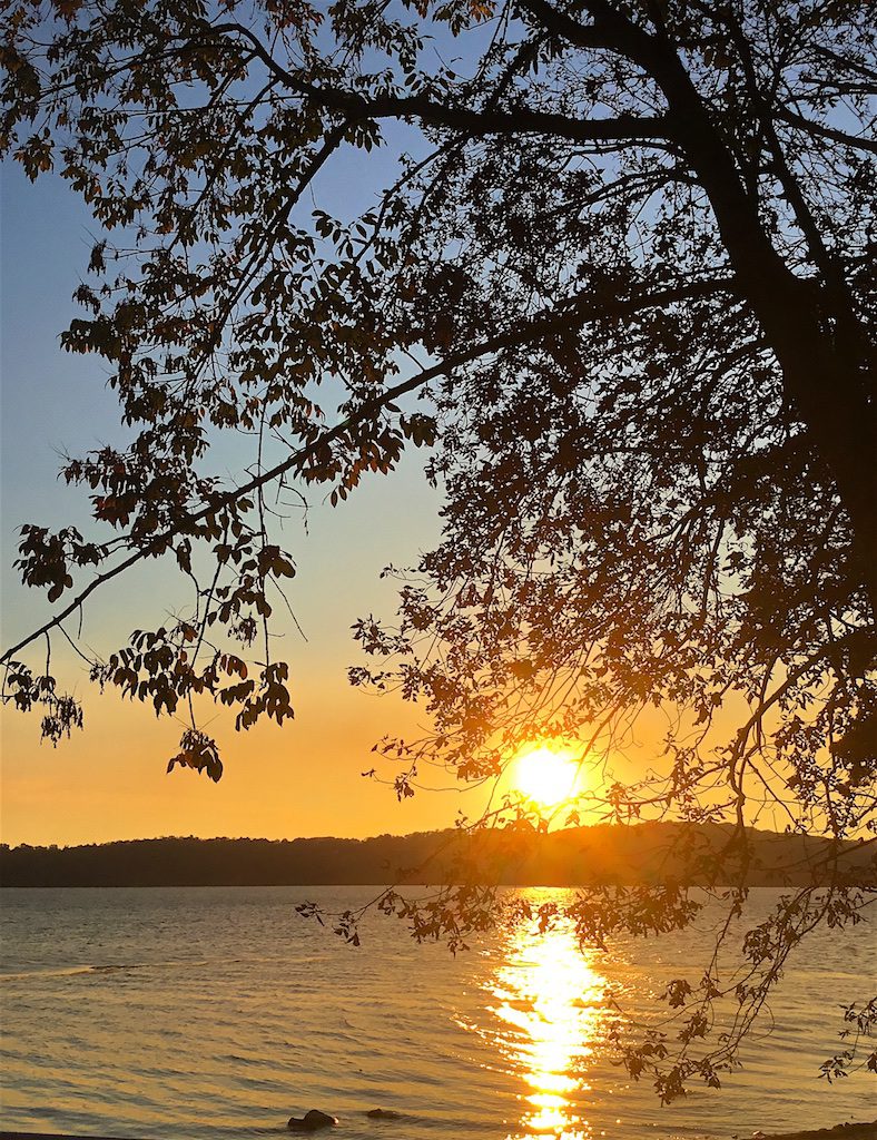 Color photograph of a sunset with a long, strong reflection on a lake. The sun appears as a bright golden ball just above a wooded ridge on the other side of the lake. The scene is viewed through branches of a deciduous tree. The sky that is bright orange over the horizon fades up to a light blue at the top of the image.