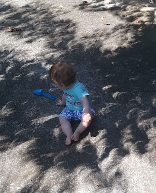 Color photograph of an infant sitting on a gray asphalt surface, surrounded by lenticular shadows from the currently still partial solar eclipse. The shadows are created by the leaves of a tree overhead (not visible in the picture). The shapes of the leaves, however, are distorted because hundreds of tiny gaps between leaves that let sun rays pass through act like pin holes of a camera obscura and project images of the sun sickle on the ground. The shade pattern created looks more like gray, irregularly overlapping stacked shingles than customary leaf shadows.