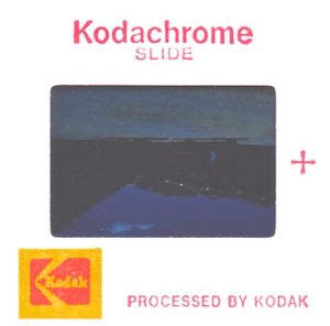 Color photograph of the back side of an original 1979 Kodachrome color slide. The red inscription at the top reads, Kodachrome SLIDE. At the bottom left is the red and yellow Kodak logo and next to it are the words PROCESSED BY KODAK. To the right of the film window is a red plus sign. The image is upside down with the emulsion facing us, as we are looking at it from the position of a projection lamp. The projection lens would be on the opposite side of the slide and invert the picture bottom to top and left to right so it will appear correctly on a reflective screen.