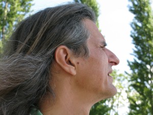 Color photograph head shot in profile view, prominently showing the artist's beautiful long hair, with trees in the background.