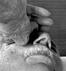Black & white close-up photograph from below the chin of a man holding his left hand against his right cheek. The bent fingers are touching the nose and appear to be crippled by arthritis. Above a short gray mustache stand out the nostrils and the eyebrow and lashes of the left eye.