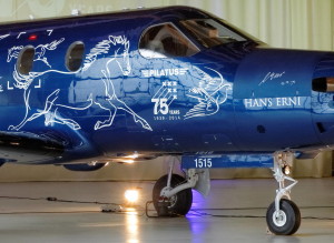 Color photograph of the cockpit section of the dark blue airplane showing the white line drawings of a small and a large running horse, the Pilatus 75 Years anniversary logo, adove, and Hans Erni's autograph.