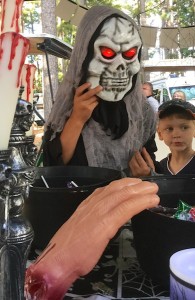 Color photograph of a tall person clad in black, with gray gauze draped over the head, shoulders, and chest, wearing a white skull mask with large, red-glowing illuminated eyeballs who is standing between large metalized gothic horror candelabra and an unmasked small boy, who appears to be clinging to the tall person for reassurance. They are looking at black buckets full of treats on a table in front of them. A very large chopped-off plastic hand rests on the rim of one of the buckets.