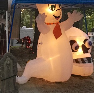 Color photograph of two large white inflatable, internally illuminated Halloween figures. One a larger than life jolly ghost with a stylized red necktie. The other, a proportionally even larger skull with big round styrofoam eyeballs, sits on the ground next to the ghost. To the right is a fake tombstone adorned by a skull, and in the background a stylized black cat, sprinkled with colorful holiday lights.