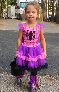 Photograph of a little girl in a ruffled pink and purple costume with a big stylized black widow spider on the chest and spider tatoo stickers on her right cheek and left upper arm. She carries a small black bucket to collect treats.