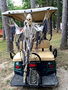 Color photograph facing a cone-headed skeleton that is attached to the back of a six-seat golf cart. It holds up its skeletonized hands and is draped in shaggy black rugs, beige gauze, and fake white spider webbing. Black plastic spiders seem to be crawling up on it from a miniature plastic headstone embossed with a skull and RIP scroll.