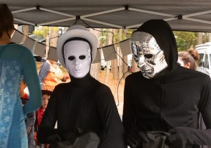 Color photograph of the chests and heads of two people holding black bags who are wearing tight fitting black jump suits with seamless gloves, hoodies, and face cover. One wears a white rimmed melon hat and shiny white mask, the other a chrome mask with embossed reticulum. There seems to be nothing behind their large eye openings; only black!
